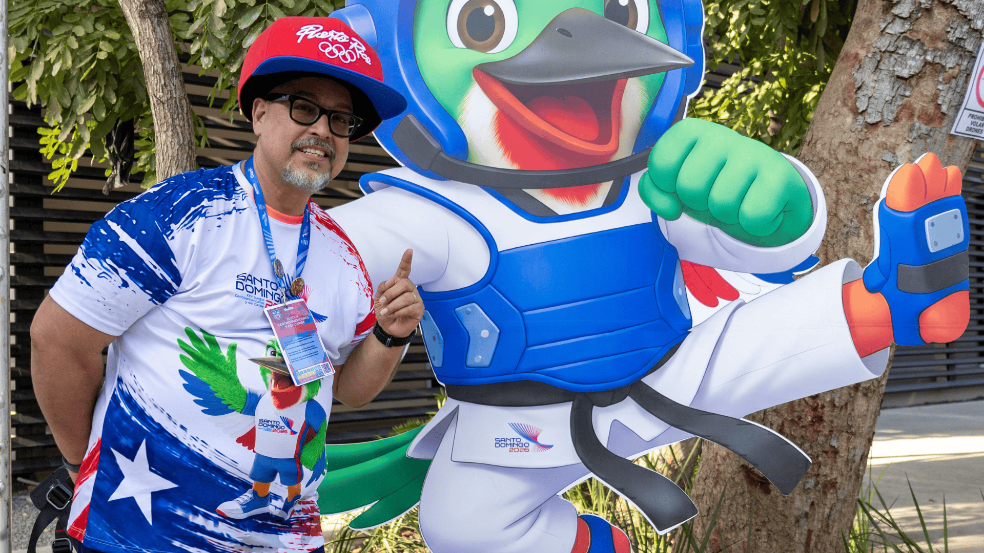Rafael posa con Colí, mascota de Santo Domingo 2026, en la entrada del pabellón de taekwondo del Centro Olímpico Juan Pablo Duarte.